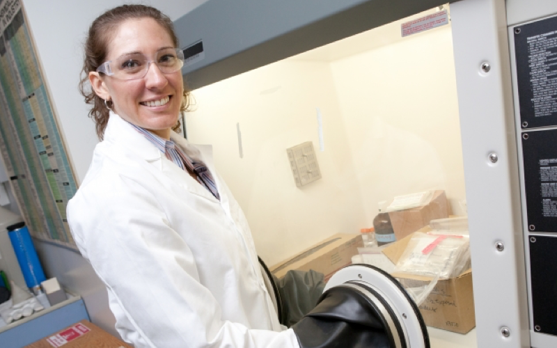 Ale Hakala smiling at the camera wearing safety goggles and a lab coat while using an anaerobic chamber in a lab