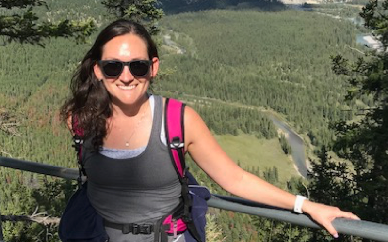 Julie Mansfield smiles at the camera. She wears sunglasses and field attire and is holding onto a rail overlooking a green landscape.