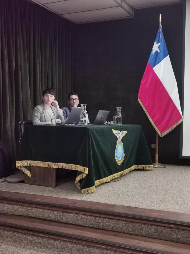 Students Mara Figueroa and Franco Sobrero facilitating English-Spanish translations during the FIG RFIP seminar. They sit at a table with a green cover and a Chilean flag stands besides them