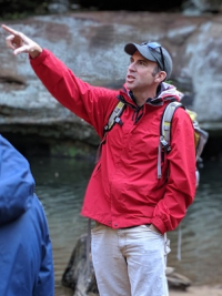 Ian Howat stands pointing in a red jacket in front of a rock formation