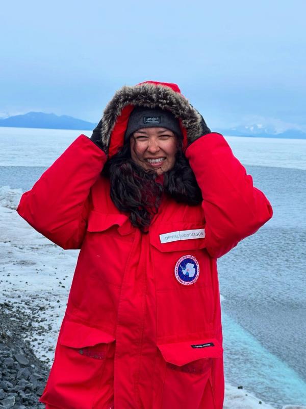 Girl with long black curly hair. Girl is wearing a big red jacker and beanie. She is smiling and holdering her jacket hood up. The background is icy and cold.