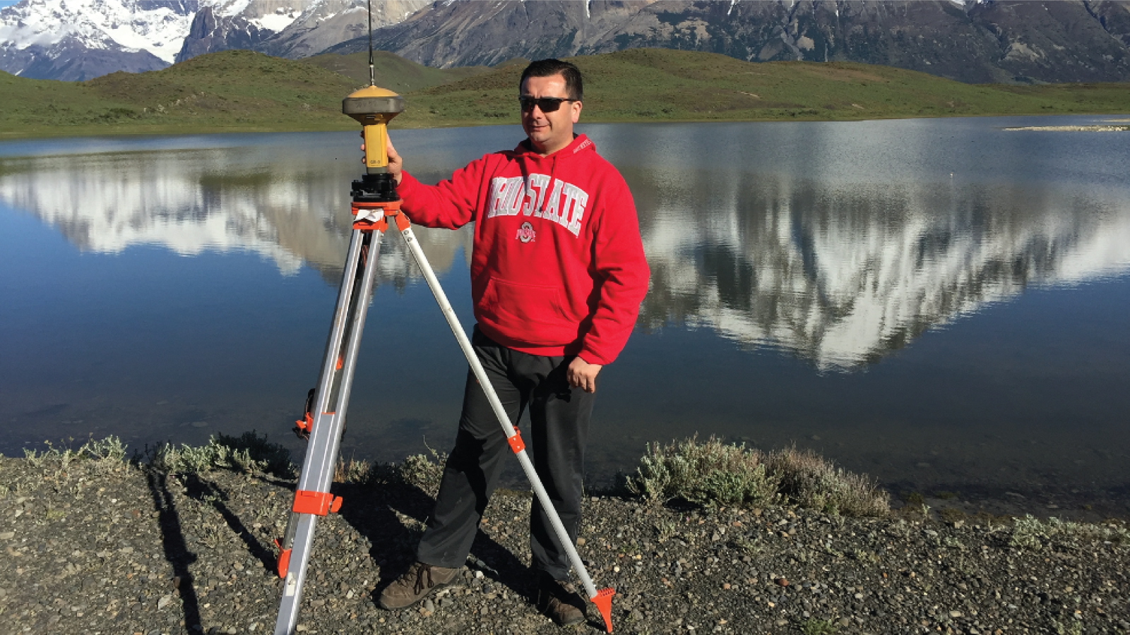 Julio carrying out field work in an OSU sweater in front of a mountain lake