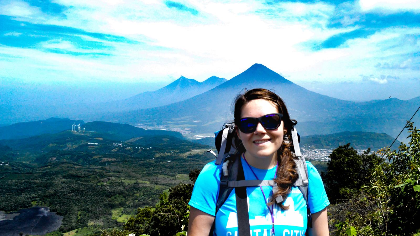 Lindsey standing on Pacaya Volcano, Guatemala, with Pacaya lava flows visible on the left, and Agua, and Fuego-Acatenango Volcanoes in the background 
